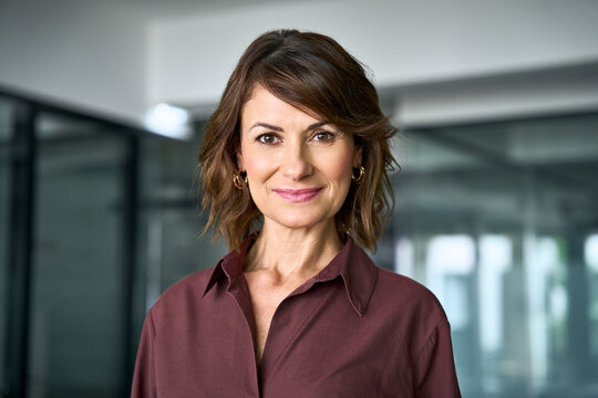 Close up face portrait of middle aged mature businesswoman in office. Attractive woman looks at camera with light smile. Confident female business director, professional in 50s poses at workplace.