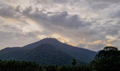 Obraz premium The sacred Velliangiri mountain peak covered in lush green forest under a dramatic stormy sky with golden sun rays breaking through heavy clouds