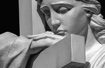 Fototapeta premium Black and white close-up of a mourning cemetery angel resting on a stone cross. A timeless symbol of grief, remembrance, faith, hope, eternal love and divine comfort.