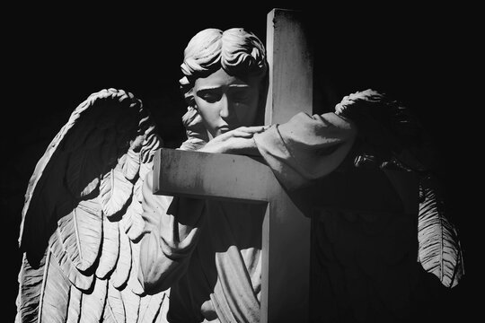 Black and white cemetery angel embracing a cross in silent prayer. Symbol of faith, remembrance, hope, eternal peace and divine protection.