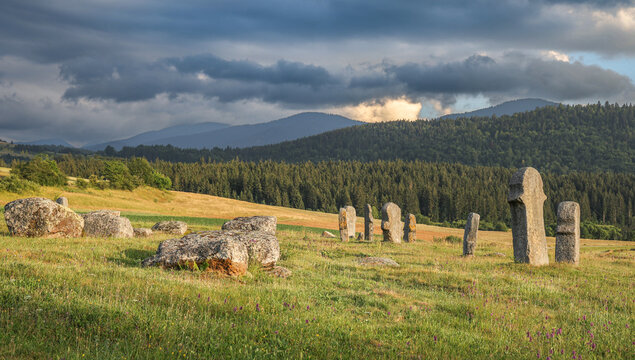 Medieval tombstones known as stecci stand in a sunlit mountain meadow beneath dramatic storm clouds, surrounded by forested hills near Novi Travnik, Bosnia and Herzegovina.