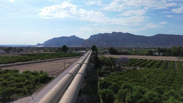 aerial view of large metal water transport pipelines running through rural agricultural landscape with green fruit orchards and distant mountains under blue horizon in scenic countryside development p