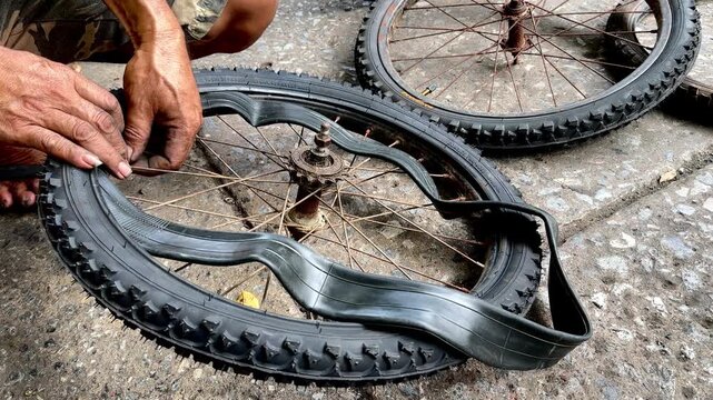 A close-up of a person's hand holding a heavily damaged bicycle tire with a large tear or hole in the sidewall. Showing a flat tire needing repair or replacement on a concrete ground.