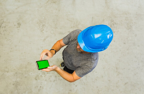 Top view of a worker in a blue hard hat using a smartphone with a green screen on a concrete floor