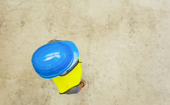 Top view of a worker in a blue hard hat and yellow safety vest walking on a concrete floor with copy space