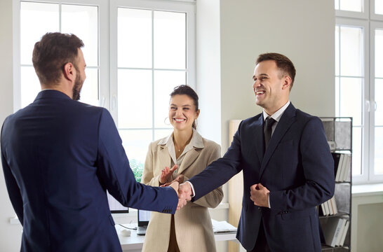 Business handshake in office meeting signals agreement. Two partners in suits seal a deal as team applauds in a bright modern workspace. Clear concept of business success and partnership.