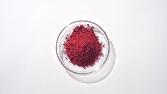 Overhead frame of a pile of red powder within a glass petri dish on a light grey background lit with orbiting light and casts moving shadow