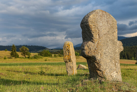 Anthropomorphic medieval stecci tombstones stand in a sunlit mountain meadow beneath dramatic storm clouds near Novi Travnik, Bosnia and Herzegovina.