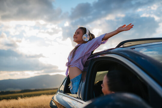 Little girl with headphones standing and leaning out of the car window during the ride.
