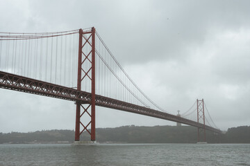 Fototapeta premium Close view of Lisbon’s 25 de Abril Bridge spanning the Tagus River, with the Cristo Rei monument visible through misty weather beneath a gray overcast sky.
