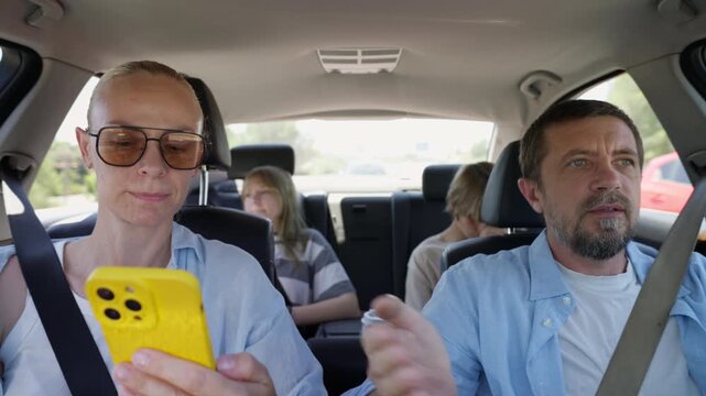 Parents debating route during family road trip while checking navigation on smartphone. Teenage son and daughter watching from back seats as family tries to find correct direction.