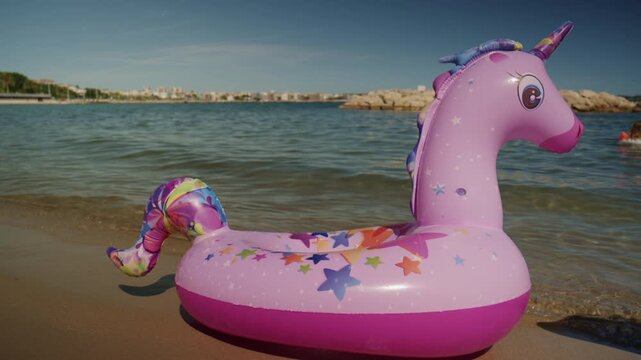 Pink unicorn inflatable float resting on the sandy shore of Plage de la Gravette in Antibes, France, with calm Mediterranean water and rocky breakwater in the background