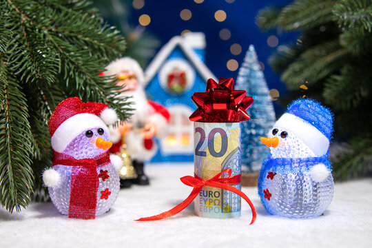A decorated euro banknote between two snowmen in colorful hats against a background of Christmas trees with Santa Claus and a blue house
