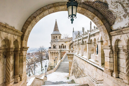 Beautiful winter view of Fisherman's Bastion at Buda Castle in Budapest, framed by a classic stone archway with a hanging lantern and snow-covered stairs.