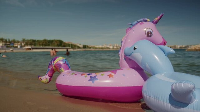 Colorful unicorn and dolphin inflatable floats on the shallow water at Plage de la Gravette with families swimming and the rocky breakwater in the background