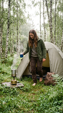 Woman camping in birch forest, holding cup near tent and camp stove with steam, outdoor adventure and travel lifestyle concept