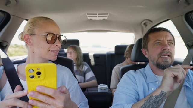 Family planning route inside modern car during road trip. Parents checking navigation on smartphone while teenage son and daughter sitting quietly in back seats, traveling through sunny countryside.