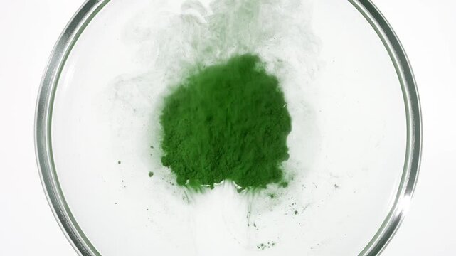 Top perspective macro shot of a foggy cloud of green dust flowing in a petri dish over a pile of powder on a simple white backdrop
