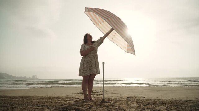 Caucasian woman in dress opens beach umbrella on sandy seashore on empty beach and sets up. Important UV protection during rest and swimming with tanning. Aesthetic shot of carefree vacation routine.
