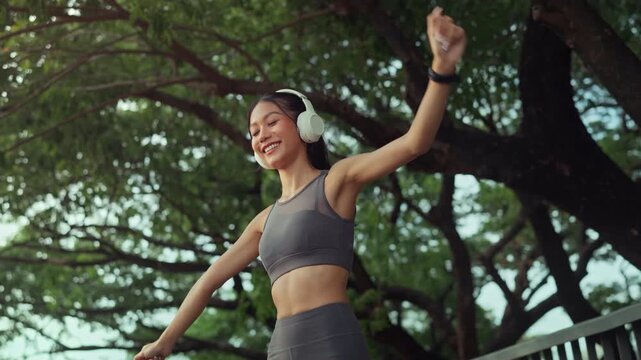 Happy Asian woman wearing headphones, smiling and dancing while enjoying music at green park, fitness female athlete relaxing after outdoor workout for active sportswear lifestyle, mental health
