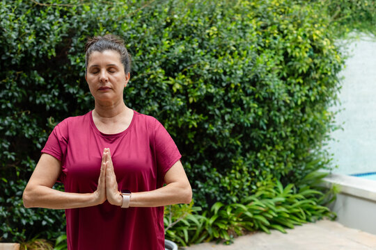 Mature female standing on patio in maroon shirt with smartwatch, pressing palms in prayer near pool