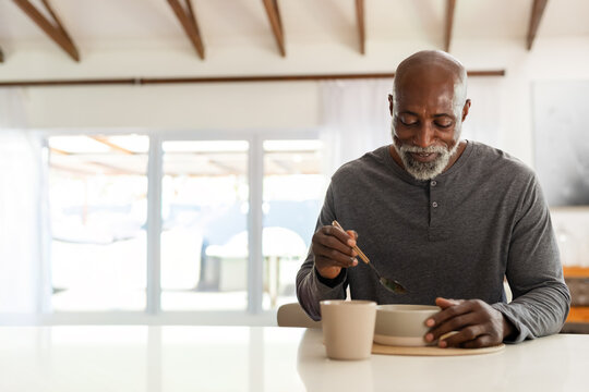 Senior African man sitting at home table wearing shirt holding ceramic bowl and spoon, copy space