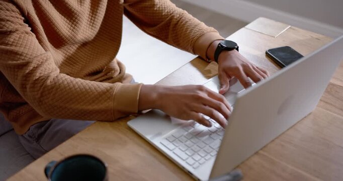 African American man wearing tan sweater typing on laptop at home, hands moving, completing work