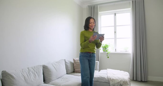 African American woman using tablet, walking to sofa at home, sitting petting calico cat to relax