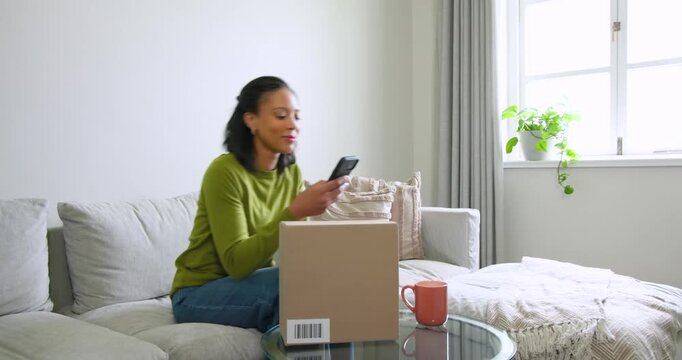 African American woman on sofa in green top checking phone on delivery alert, inspecting box label