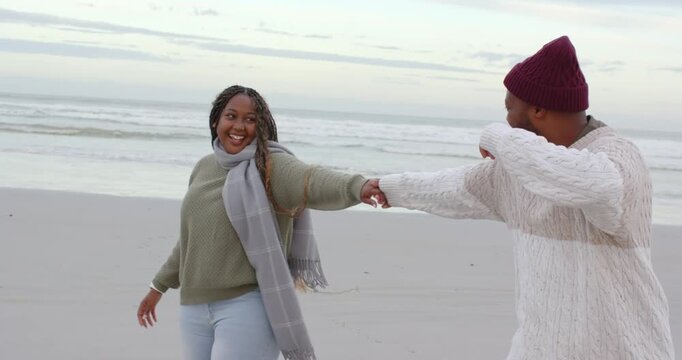 African couple walking sandy shore, man fixing beanie pulling woman twirling for fun then embracing