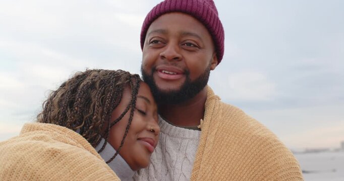 African American couple hugging in blanket at shore, chilly breeze, man wearing beanie staying warm