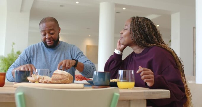 African American couple sitting at table, woman talking causing man slicing bread for breakfast
