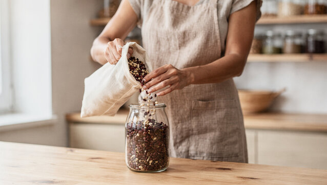 Woman pouring dried beans from a reusable cloth bag into a glass jar in a sustainable, zero-waste kitchen setting