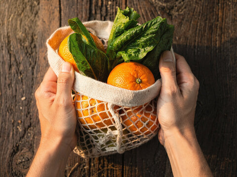 Person holding a reusable mesh bag filled with fresh oranges and green leafy vegetables over a rustic wooden background