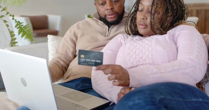 African American couple on couch, woman holding card urging man typing on laptop completing payment