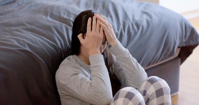 Woman sitting on floor, covering face then lowering hands, pressing head, calming by bed in pajamas