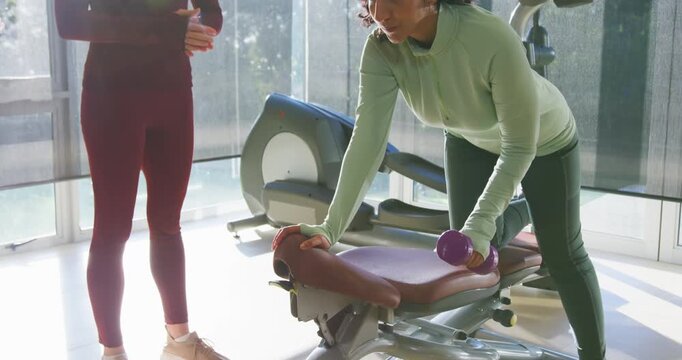 Female coach cueing trainee in green kneeling on bench pulling purple weight for strength in gym