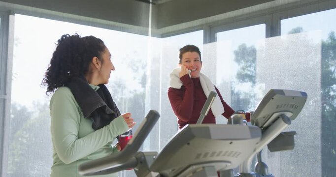 Diverse female friends cooling down on treadmills chatting dabbing with towel and sipping bottle