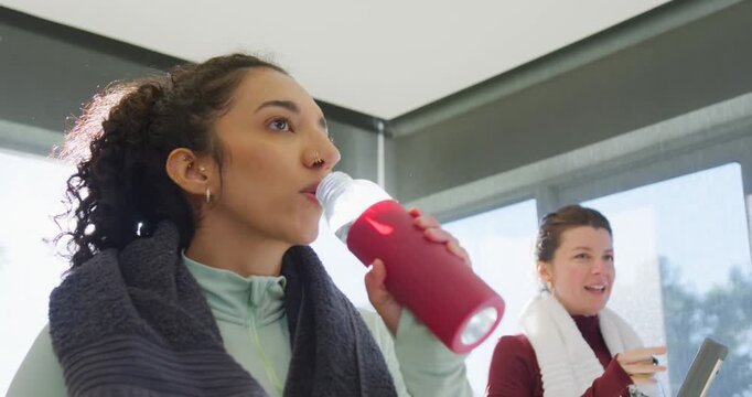 Diverse female duo rehydrating post-workout biting apple and sipping red bottle in gym, copy space