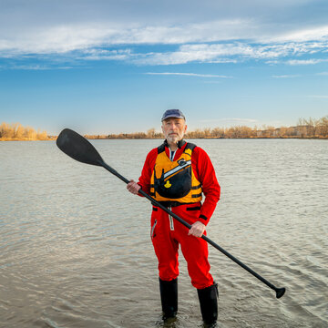 environmental portrait of a senior male paddler in a drysuit and life jacket holding stand up paddle on a shore of a lake