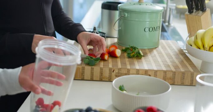 Diverse women friends trimming strawberry stems on board, adding yogurt to blender cup for smoothie