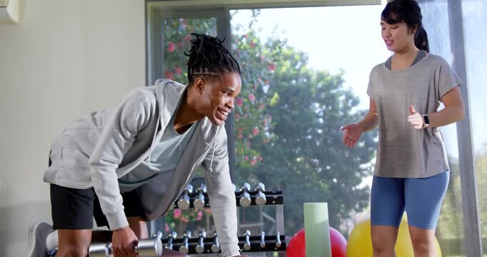 African man trainee following Asian trainer cueing, picking dumbbell and doing rows in gym for form