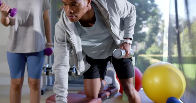 African American man adjusting bench rowing dumbbell as Asian woman curling weights training at gym