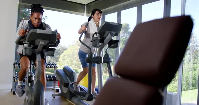 Gym partners pedaling bike and striding elliptical in gym after mounting, doing cardio with towel