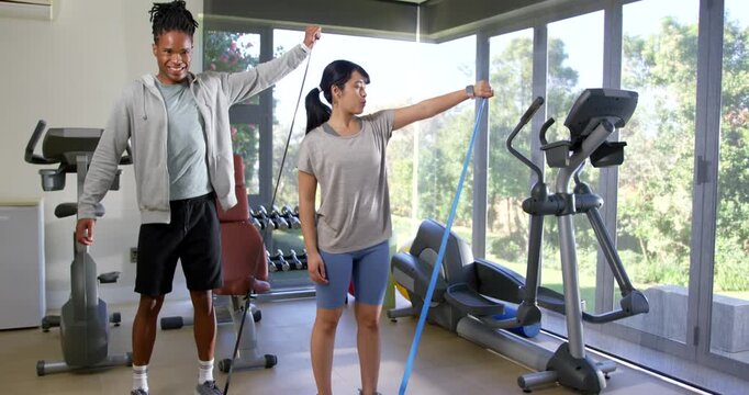 African American trainer raising arm, guiding trainee holding blue resistance band at gym for form