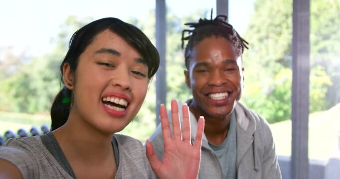 Diverse couple in gray responding to camera prompt, speaking and waving on sunroom bench by weights