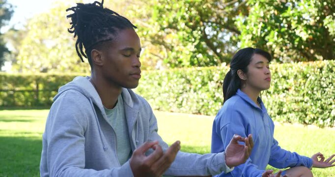 African American man and Asian woman meditating on grassy lawn in park, closing eyes, forming mudra