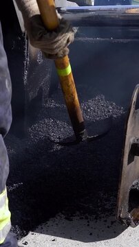 Worker Loading Shovel From Asphalt Paver Hopper Slow Motion. Road construction worker wearing boots and gloves scoops hot asphalt with a shovel near machine hopper wings during street resurfacing.