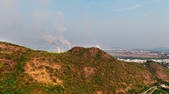 Aerial over a dry scrub hilltop with distant industrial smokestacks on the horizon
