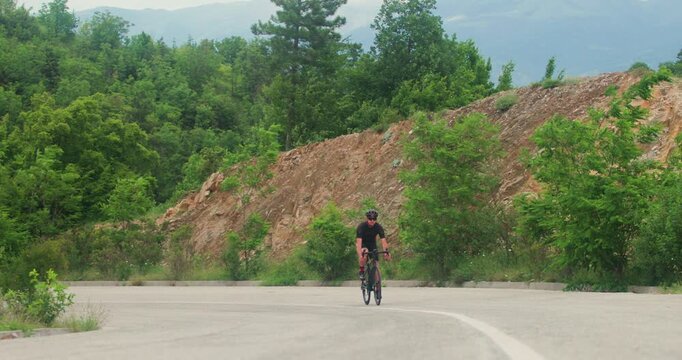 A male cyclist in a black and red cycling suit and helmet rides his road bicycle uphill on a winding mountain road, training for an endurance race in beautiful natural surroundings.